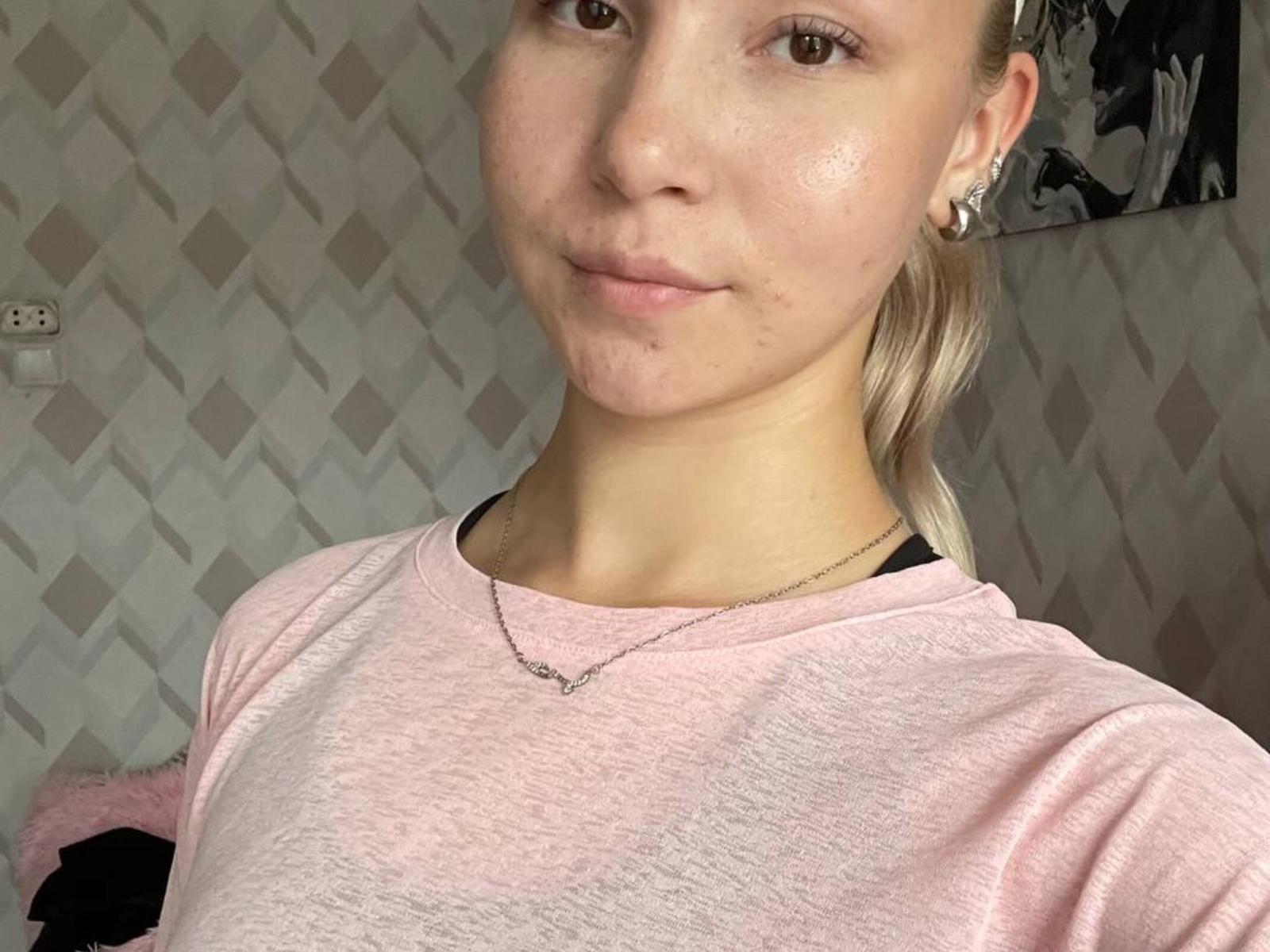 Marguerite Geppert tiene una ligera sonrisa, piel clara y algunos granitos pequeños en las mejillas. Lleva una camisa rosa, un collar delgado, y su cabello rubio está recogido..