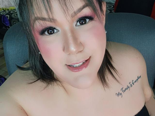 GiiannaMorrys with pink eye makeup, black hair, nose ring, smiling, showing off her tits, tattoo on right shoulder, sitting in gray chair, looking at camera.