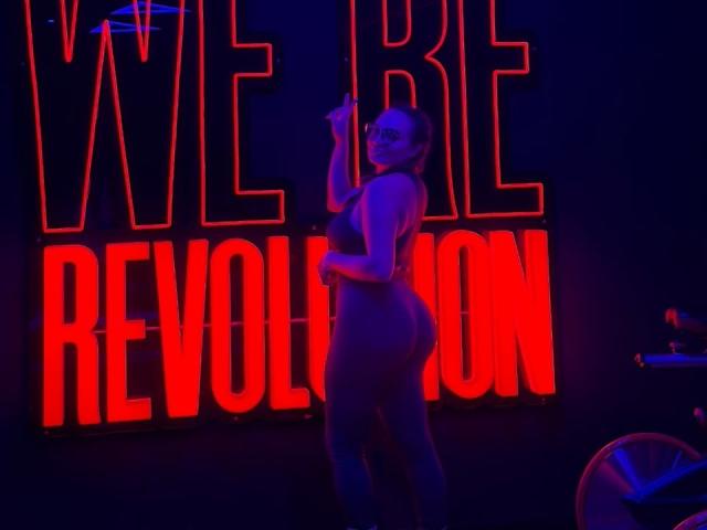 GalaMalkova in a neon-lit club, wearing a tight, shiny, black bodysuit, with her back to the camera, showing her round ass, holding a cigarette, in a sultry pose.