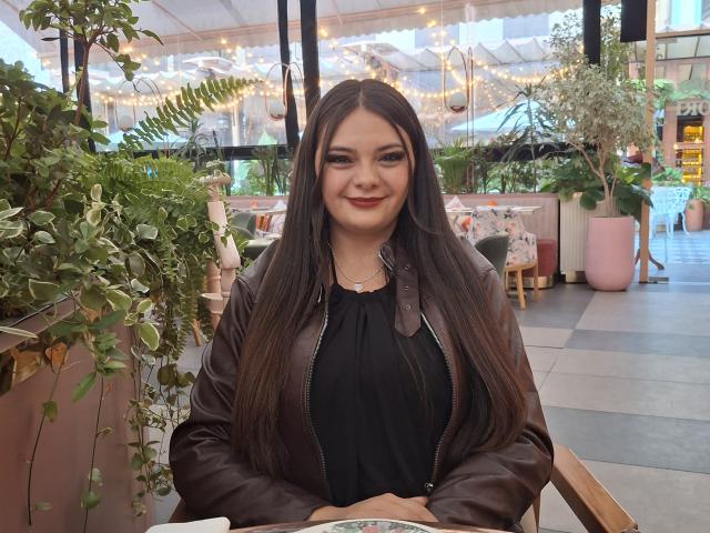 KatherinLatin, com cabelos castanhos longos, vestindo uma blusa preta e jaqueta de couro marrom, sentada em um café, sorrindo para a câmera, segurando uma xícara branca, com os seios à mostra..