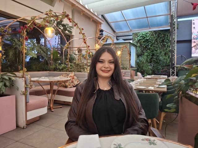 Katherin Latin a de longs cheveux noirs, porte un haut noir avec une veste marron, et sourit à l'appareil photo avec du rouge à lèvres. Elle est assise à une table dans un café confortable, rempli de plantes..
