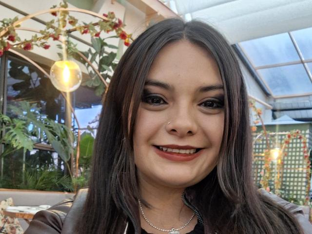 Katherin, met rechtbruin haar, zwarte eyeliner, rode lipstick, een klein neuspiercing, lachend, draagt een bruine leren jas en een zilveren ketting..