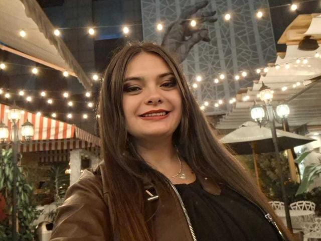 Katherin Latin a de longs cheveux bruns, un piercing au nez, et elle sourit avec du rouge à lèvres. Elle porte une veste marron et un haut noir, ainsi qu'un collier..