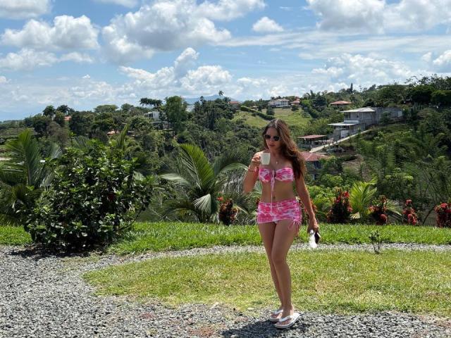 Gabrielafoxxy, portant un bikini floral rose, tenant une glace, debout sur du gravier, chaussée de baskets blanches, portant des lunettes de soleil et ayant de longs cheveux bruns, dans un paysage ensoleillé, verdoyant et vallonné..