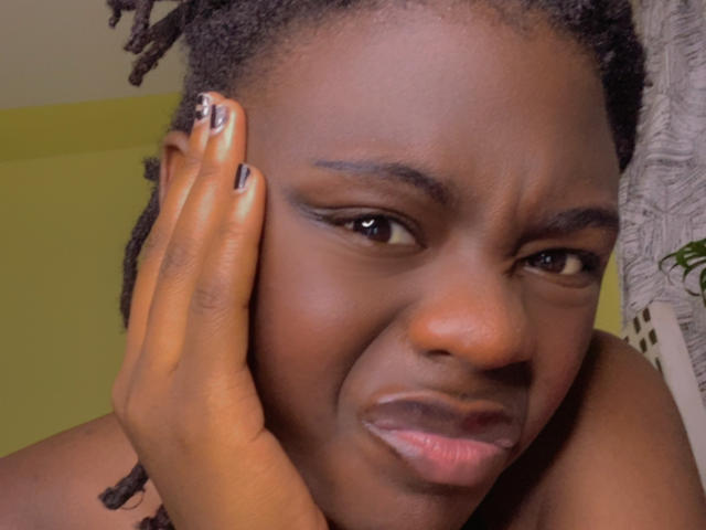 BustySabrina a la peau foncée, des dreadlocks de longueur moyenne, et une expression pensive, avec sa main sur sa joue, montrant du vernis à ongles noir avec des points blancs. Ses lèvres sont légèrement entrouvertes..