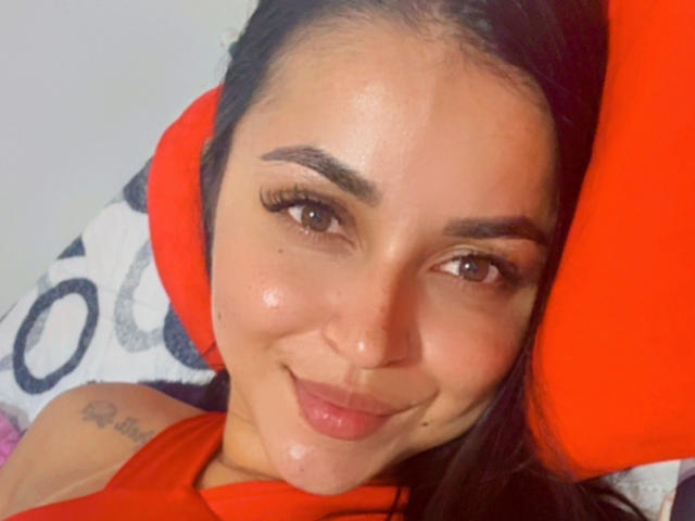 HikariChloe, con una sonrisa encantadora, cejas oscuras, pestañas largas y un tatuaje en el hombro, acostada sobre un cojín rojo con un ligero brillo en su rostro..