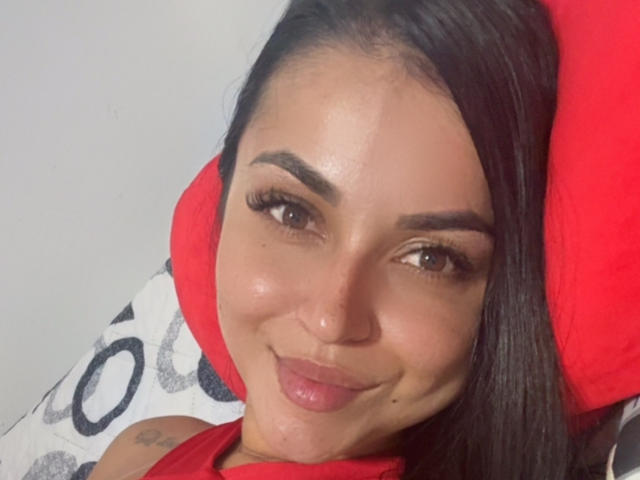 HikariChloe, avec ses yeux marron brillants, ses sourcils épais et ses longs cheveux noirs, souriant avec du rouge à lèvres, portant une chemise rouge, allongée sur un oreiller rouge et un oreiller blanc avec des points noirs..