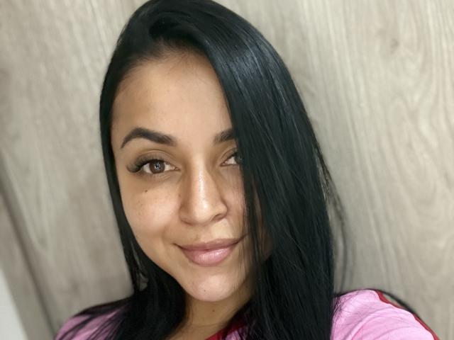 HikariChloe, com cabelos pretos longos, pele morena, olhos castanhos e um sorriso discreto, vestindo uma camisa rosa, parece adorável com seus cílios e nariz pequeno..