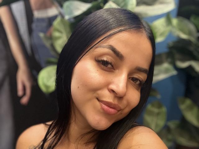 HikariChloe, con cabello negro, ojos marrones y una pequeña marca de nacimiento en su mejilla izquierda, sonríe suavemente a la cámara. Sus hombros desnudos son visibles..
