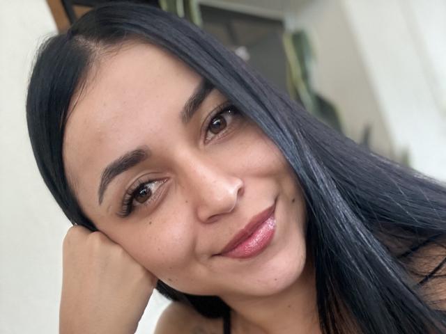 HikariChloe tem cabelos pretos longos, olhos castanhos e um nariz pequeno. Ela está sorrindo, usando batom rosa, e sua mão está tocando sua bochecha. Sua pele é clara e lisa..