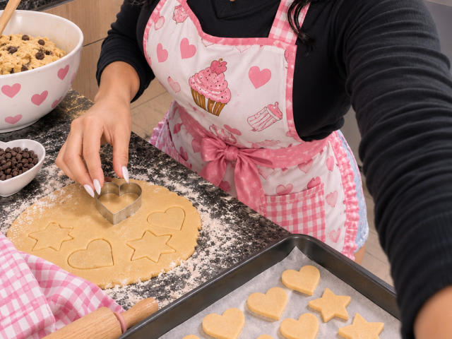 NenaBlack con delantal rosa con forma de corazón, camisa negra de manga larga, cortando masa para galletas con forma de corazón y estrella con un cortador de metal sobre una encimera de granito negro. Rodillo de madera y tela rosa a cuadros..