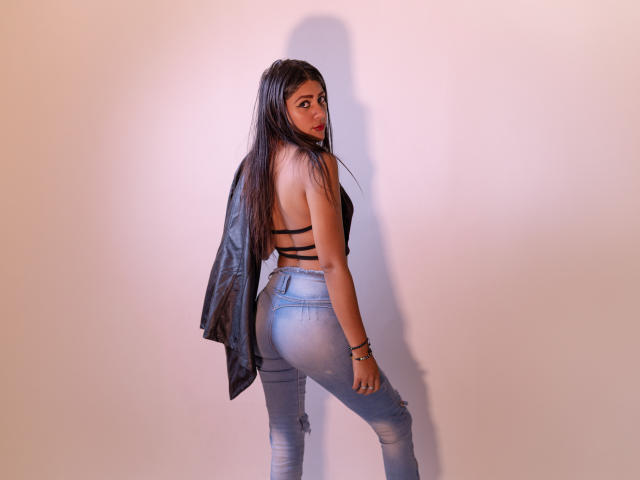 NataliaCruz with long black hair, back to camera, wearing a black strappy top, blue jeans, and a black leather jacket, showing her ass and looking over her shoulder.