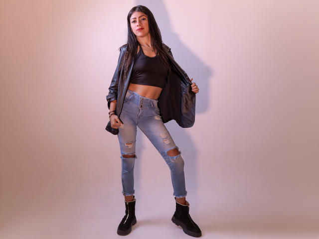 NataliaCruz with long dark hair, black crop top, black leather jacket, ripped blue jeans, and black boots, standing confidently with a slight smile, hands on jacket.