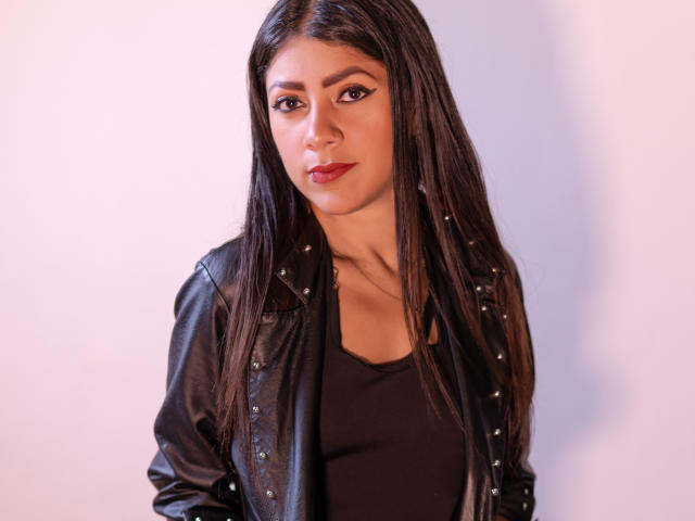 Natalia Cruz tiene el cabello largo y oscuro, lleva una chaqueta de cuero negra con tachuelas y una blusa negra, con lápiz labial rojo, y una expresión seria, mirando directamente a la cámara..