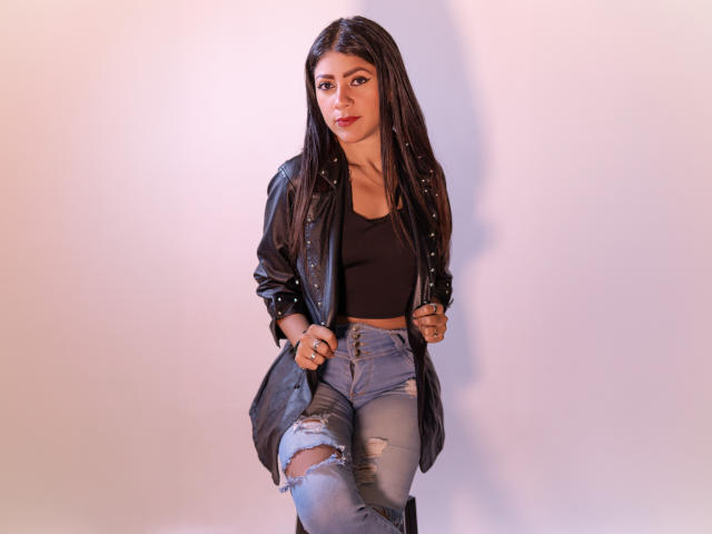 Natalia Cruz, com longos cabelos pretos, vestindo uma jaqueta de couro preta, um cropped preto e jeans azuis rasgados, está em pé com uma expressão confiante e ligeiramente sedutora, com as mãos nos jeans..