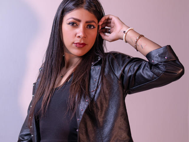 Natalia Cruz, com cabelos longos e escuros, vestindo uma jaqueta de couro preta e uma blusa preta, tem uma expressão séria, toca sua orelha com uma pulseira de ouro e tem uma tatuagem no pulso..