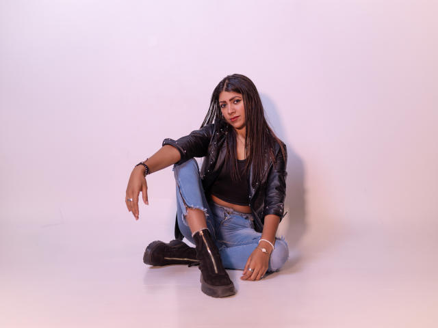 Natalia Cruz, com cabelos longos e pretos, jaqueta de couro preta, cropped preto, jeans rasgados e botas pretas, sentada no chão com uma expressão relaxada e ligeiramente sedutora..