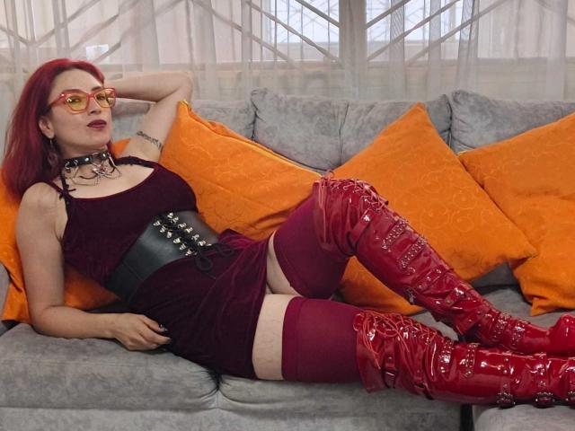 QueenScarletSecret con capelli rossi, occhiali, collana nera, vestito color borgogna, corpetto nero, stivali rossi alti fino alla coscia, sdraiata su un divano grigio con cuscini arancioni, mostrando il sedere..