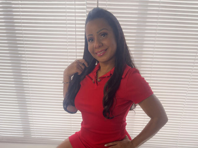 AlondraMonique mit langen, dunklen Haaren, trägt ein rotes Poloshirt, lächelt, eine Hand am Hüfte, die andere Hand im Haar, Licht von Jalousien im Hintergrund, steht, zeigt kleine Brüste, süß, sexy..