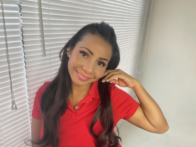AlondraMonique hat langes, dunkles Haar, braune Augen und trägt ein rotes Poloshirt. Sie lächelt mit pinkfarbener Lippenstift, berührt ihr Haar und trägt kleine Ohrringe. Ihr Hintern ist nicht sichtbar..