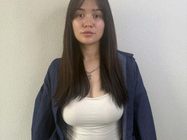 Anna Bauknight, avec de longs cheveux bruns, chemise bleue portée en dehors des épaules, débardeur blanc, petite poitrine, expression neutre, collier en argent, debout devant un mur blanc texturé, regardant directement la caméra..