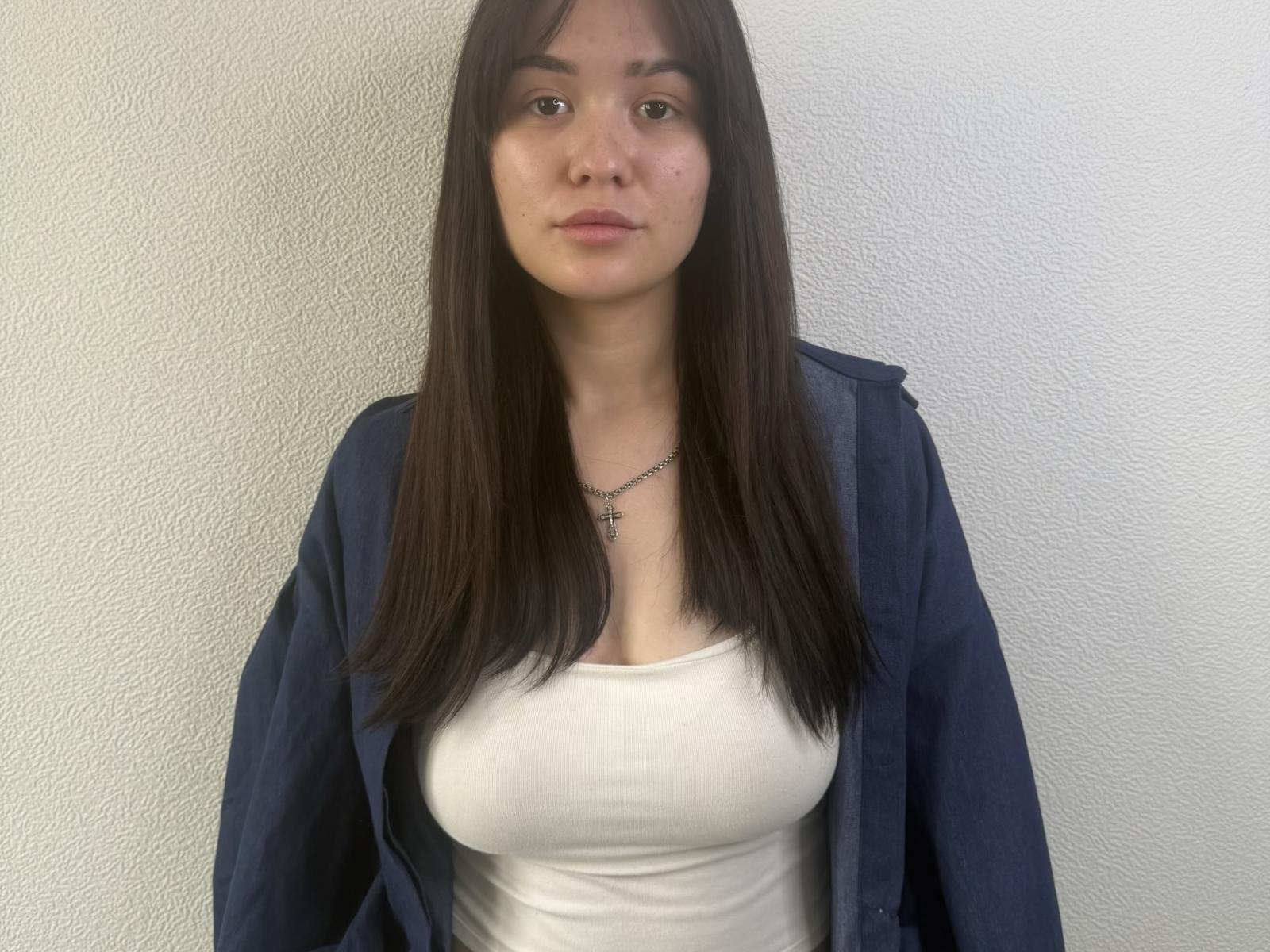 Anna Bauknight, avec de longs cheveux bruns, chemise bleue portée en dehors des épaules, débardeur blanc, petite poitrine, expression neutre, collier en argent, debout devant un mur blanc texturé, regardant directement la caméra..