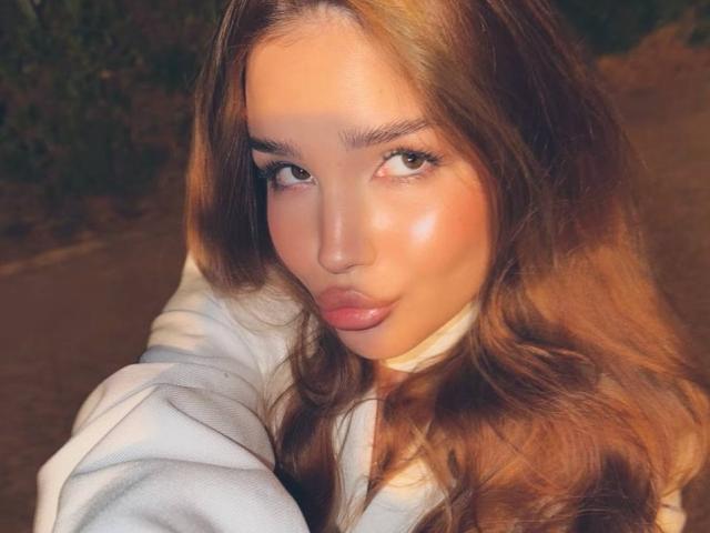 GenieCanup's pouty face, brown wavy hair, and glossy pink lips are framed by her white shirt. Her eyes are half-lidded and she's giving a subtle, sexy look.