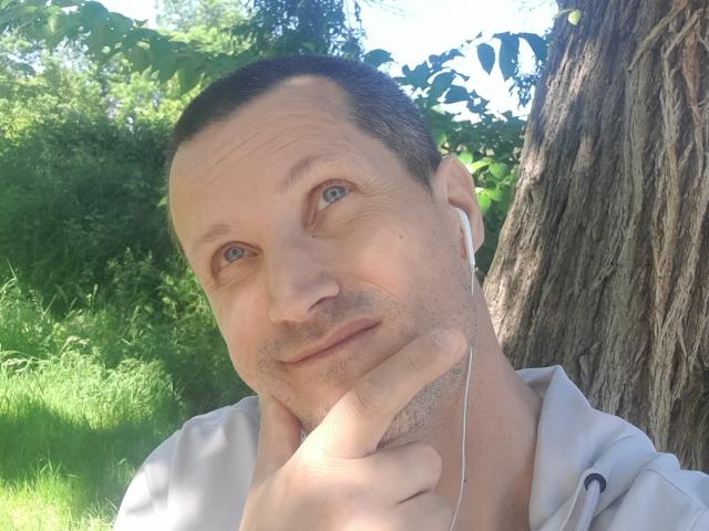MisterFredX with short brown hair, light blue eyes, and slight stubble, touching his chin with a finger, wearing white earbuds, and a light shirt, standing near a tree.