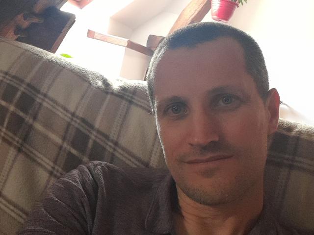 MisterFredX ha i capelli castano corti, un leggero baffo, e indossa una camicia marrone scuro, seduto su un divano a quadri con un sorriso appena accennato..