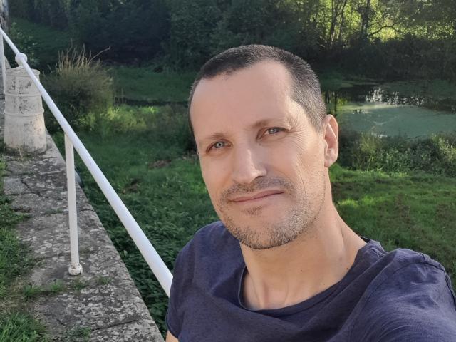MisterFredX a la tête rasée, des yeux bleu clair et un léger sourire, il porte une chemise violette, avec une rampe blanche sur sa gauche et de la verdure derrière lui..