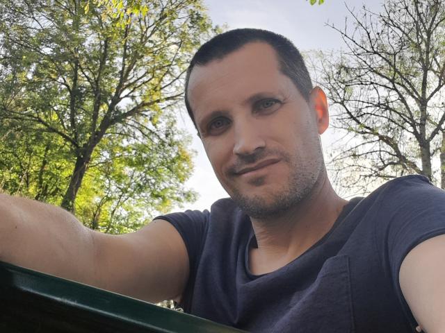 MisterFredX, com cabelo curto e escuro, barba rala e camiseta azul escura, apoiado em um corrimão de metal verde, olhando para baixo com um leve sorriso, fundo externo, luz do dia, sombras de árvores..