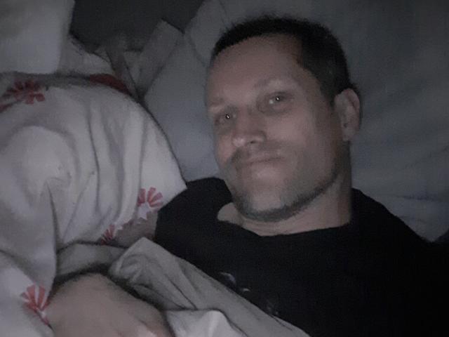 MisterFredX, een kale, middelbare leeftijd man met een korte baard, ligt in bed, gekleed in een zwart shirt, met een lichtelijk zelfvoldane blik, en een deken met rode patronen..