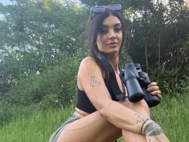 AnacondaX met tatoeages, een zwarte sportbeha en bruine shorts, die verrekijkers vasthoudt, zittend op het gras, donker haar, zonnebril op haar hoofd, bos op de achtergrond, zelfverzekerde glimlach, zichtbaar achterwerk..