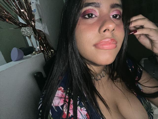 SiennaTaylor con trucco rosa per gli occhi, lunghi capelli neri, labbra rosa carnose, top floreale che mette in risalto un seno abbondante, collana choker, unghie rosse, che guarda la fotocamera, con una mano vicino all'orecchio..