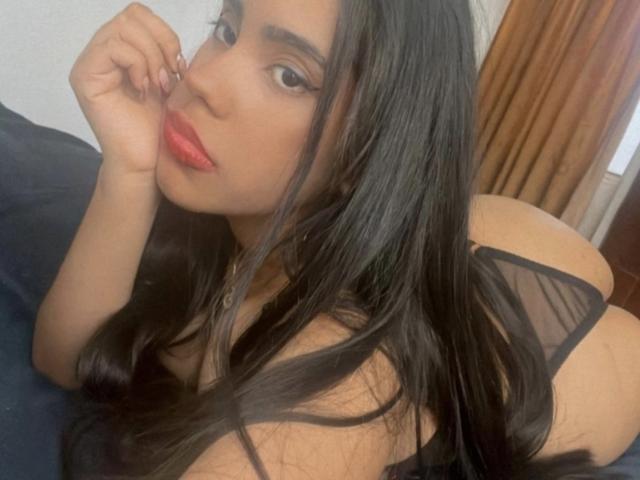 SiennaTaylor with long black hair, red lipstick, lying on a bed in black sheer lingerie, showing her ass and looking at the camera with a hand near her face.