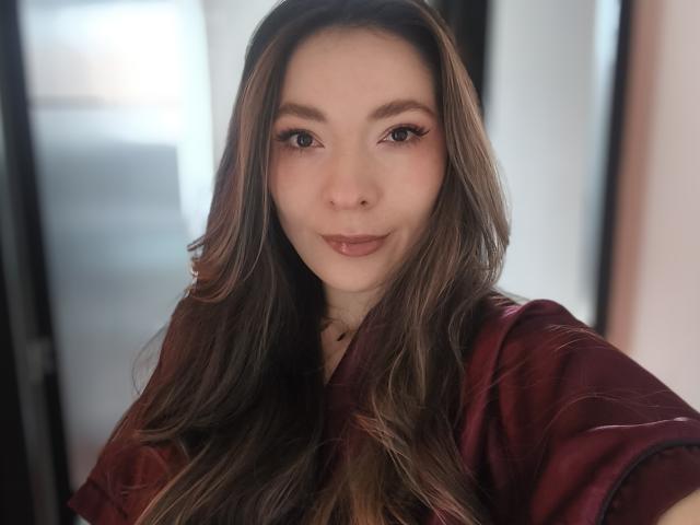 TaniaSandersX, met lang bruin haar, draagt een rode blouse en heeft een lichte glimlach en bruine ogen. Haar make-up omvat lange wimpers en subtiele roze lippenstift..