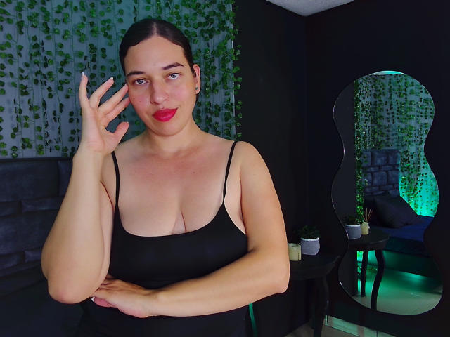 Rachel Wilson mit rotem Lippenstift, schwarzem Tanktop, das ihre Brüste zeigt, Hand in der Nähe des Gesichts, braunes Haar, schwarzer Hintergrund, Spiegelung einer grünen Pflanze, Spiegel, dunkler Raum, ruhiger Gesichtsausdruck, leichte Röte..