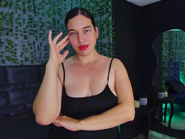 Rachel Wilson, avec du rouge à lèvres, les cheveux foncés attachés en queue de cheval, portant un débardeur noir qui dévoile sa poitrine, levant un doigt, debout dans une pièce sombre avec des plantes vertes en décoration..