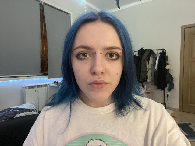 NoreneLournes, con cabello azul, ojos marrones y perforaciones faciales, vestida con una camisa blanca con un estampado de dibujos animados, mirando directamente a la cámara. Habitación con ropa en un perchero..
