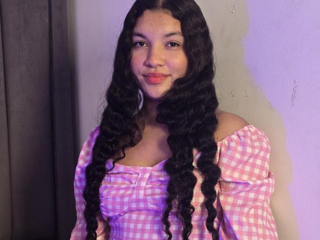 BigAssmilu heeft lang, krullend zwart haar, een roze geruit top met een open schouder, en roze lippenstift, ze glimlacht met kleine borsten, en staat tegen een effen muur, met een grijze gordijn..