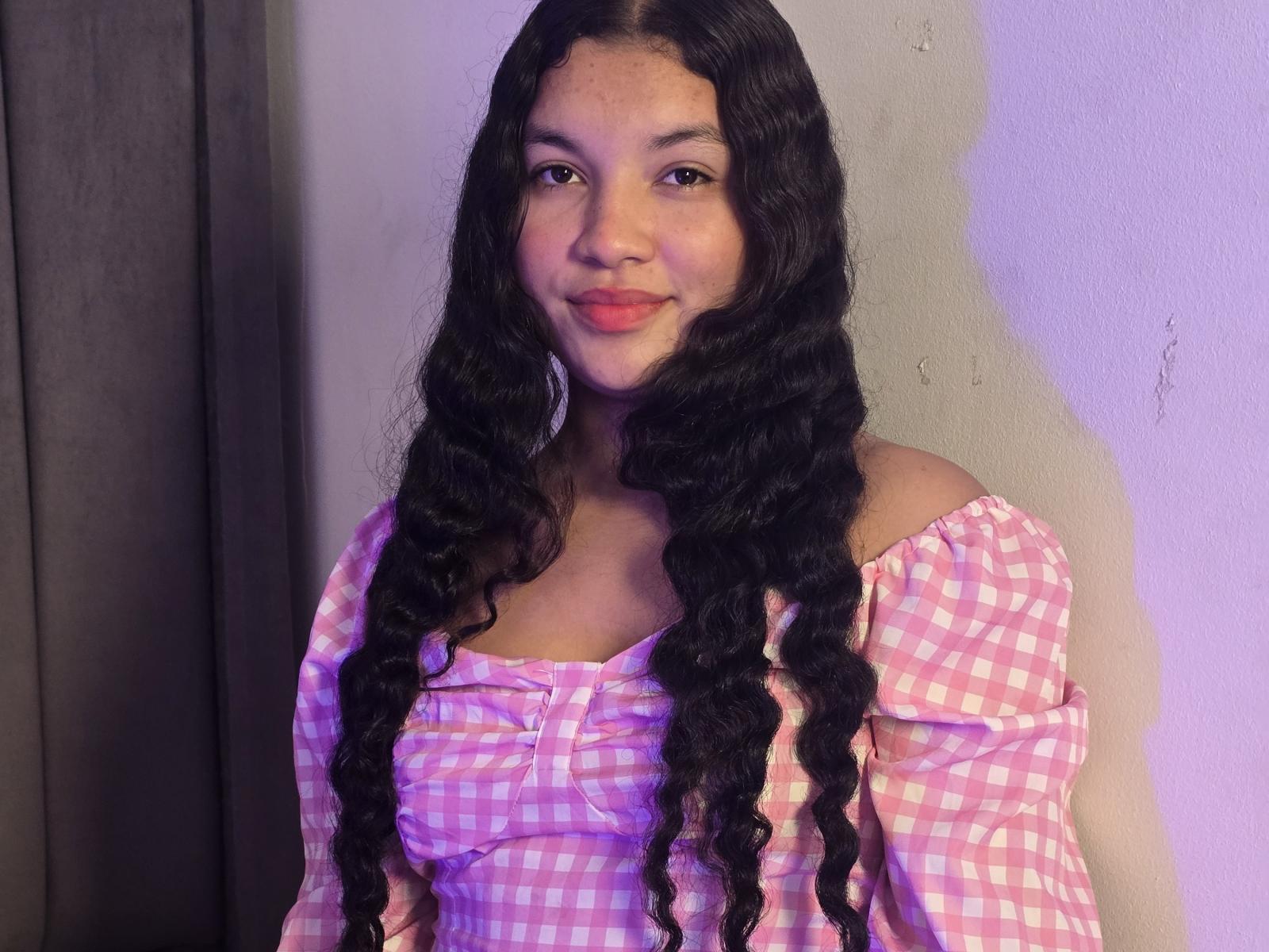 BigAssmilu ha lunghi capelli neri e ricci, una maglietta rosa a quadretti con una spalla scoperta e rossetto rosa, sorride con piccoli seni, ed è in piedi contro un muro spoglio, con una tenda grigia..