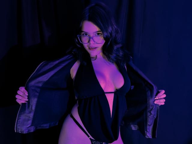 AlexaMissFortune with glasses, dark wavy hair, black lingerie, and a black jacket open, revealing her large boobs and a thong, standing in purple-blue lighting.