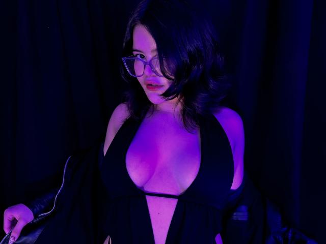 AlexaMissFortune with big boobs in a black, deep-cut dress, wearing glasses, black hair, and a black jacket, against a dark blue-purple light.