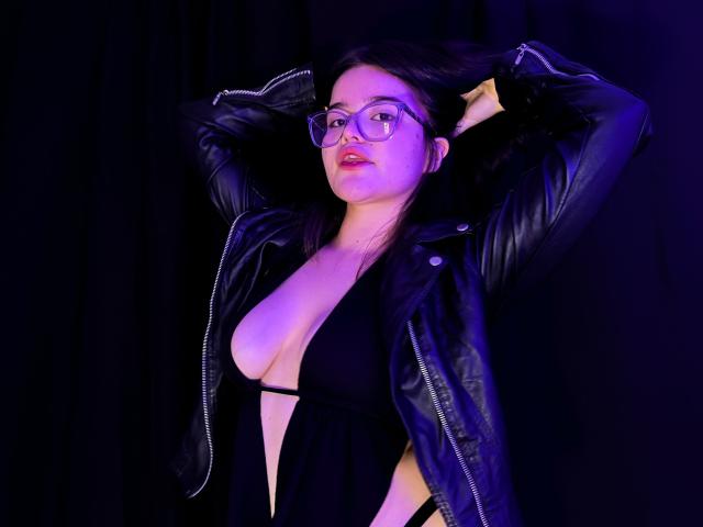 AlexaMissFortune with glasses, black leather jacket, and deep black dress, showing her big tits in a low-cut top, arms raised, dark background, purple lighting.