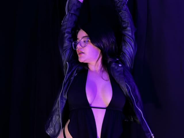 AlexaMissFortune with glasses, black dress, deep cleavage, black leather jacket, arms raised, purple lighting, dark background, sexy, looking to the side, black hair, confident.