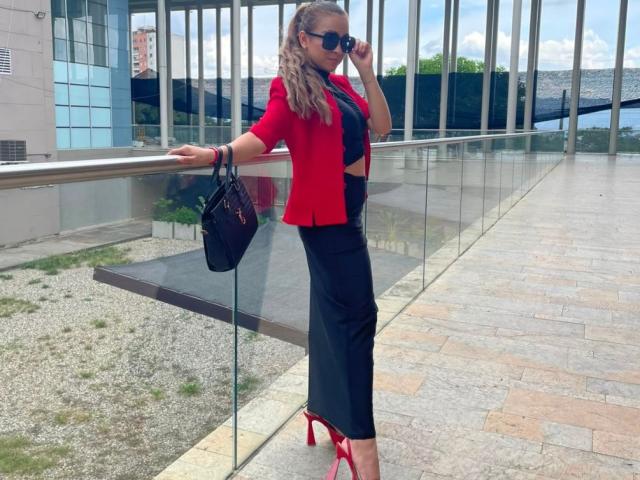 AriannaBlake with long blonde curls, red jacket, black pants, red heels, black handbag, and sunglasses, leaning on glass railing, showing her tits and ass, outside on a stone path.