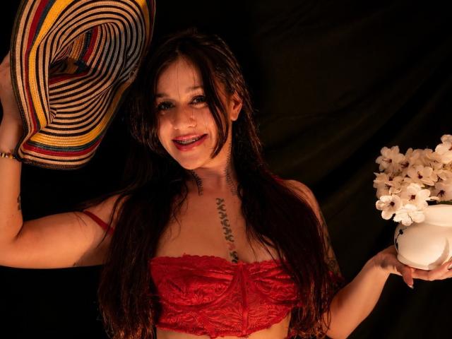 Abby Montgomery, com cabelo castanho molhado, sutiã de renda vermelho, segurando um chapéu colorido, um vaso de flores branco, sorrindo, com tatuagem no peito, fundo escuro. Seios, sutiã vermelho, textura de renda, expressão feliz..