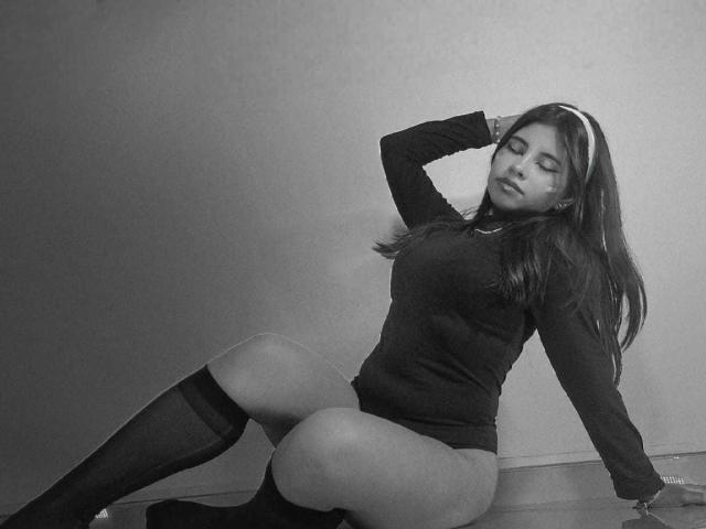 CelesteMorel est assise par terre, vêtue d'un haut noir à manches longues et de chaussettes montantes, avec une main dans ses cheveux, montrant ses seins et ses fesses. Photo en noir et blanc..
