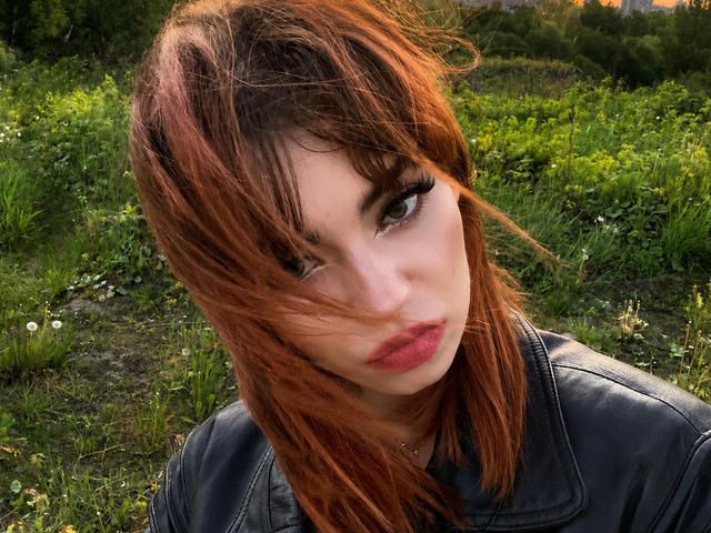 YettaGlumac, avec ses cheveux roux, clignant de l'œil et faisant la moue, portant une veste en cuir noir, ses seins ne sont pas visibles, debout dans un champ herbeux, avec un léger vent dans ses cheveux..