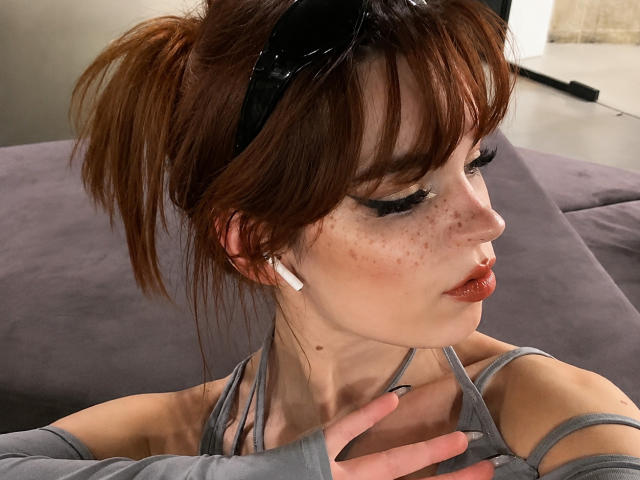 YettaGlumac mit Sommersprossen, rotem Lippenstift, braunen Haaren im Pferdeschwanz, schwarzen Sonnenbrillen, einem grauen Oberteil, weißen Kopfhörern und Handschuhen ohne Finger, die zur Seite schaut. Brüste sind unter dem Oberteil sichtbar..